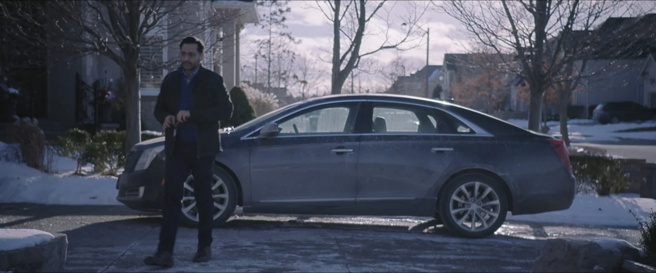 The Lie (2018) movie still: fullbody — A suburban residential driveway features a modern dark sedan parked on paved asphalt. A man in winter…