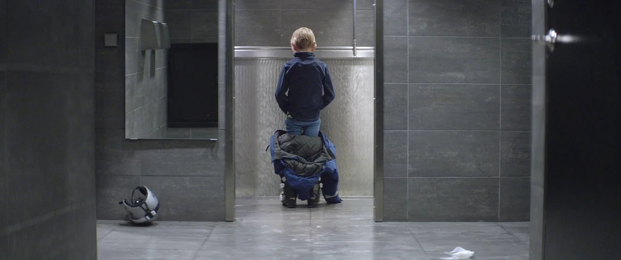 Force Majeure (2014) movie still: fullbody — A modern public restroom with grey tile walls and a long stainless steel trough. A young boy stands at…
