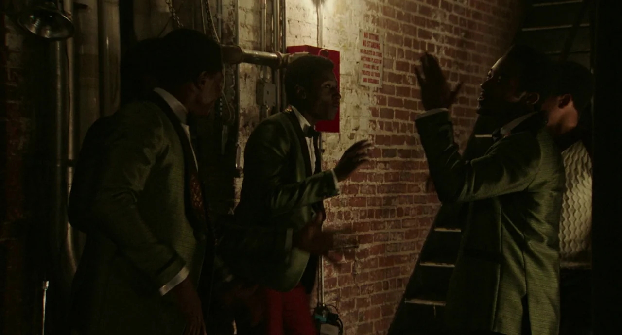 Detroit (2017) movie still: medium — A dimly lit industrial hallway features exposed brickwork, metallic pipes, and a mounted notice sign. Three men…