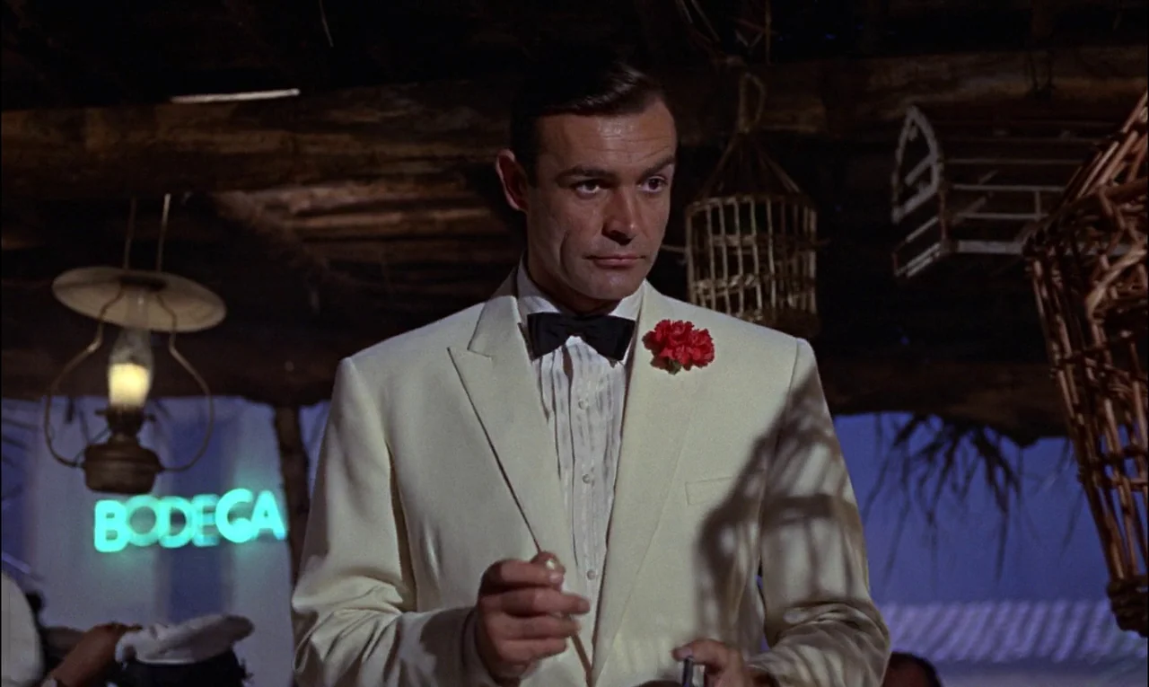Goldfinger (1964) movie still: medium — A dimly lit establishment features wooden rafters and suspended woven cages under a roof. A man in a formal…