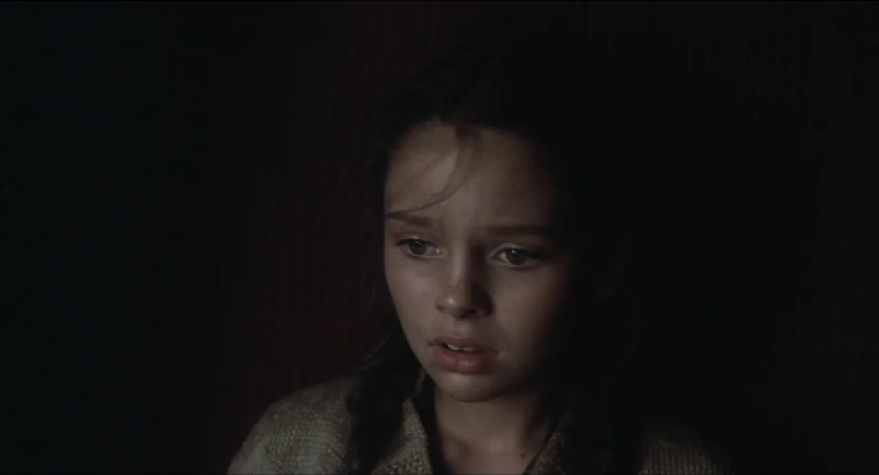 Sentimental Value (2025) movie still: closeup — A dark, minimally lit interior space with shadows obscuring the surroundings. A young girl with…