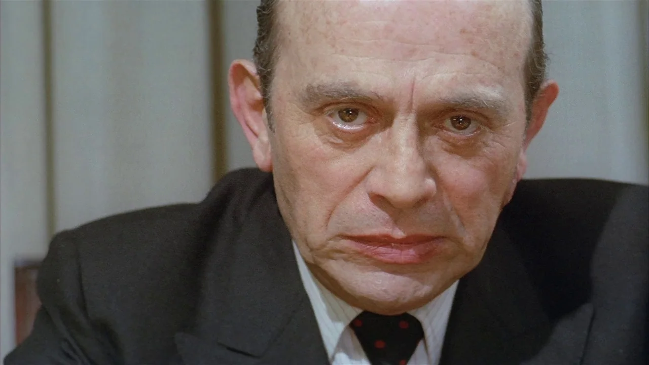 Death Occurred Last Night (1970) movie still: closeup — An older man wearing a suit jacket and tie faces the camera directly. The background…