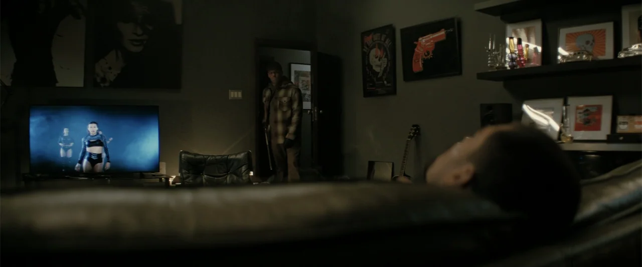 A Working Man (2025) movie still: medium — A dark apartment living room features a person lounging on a leather sofa. A man holding a baseball bat…