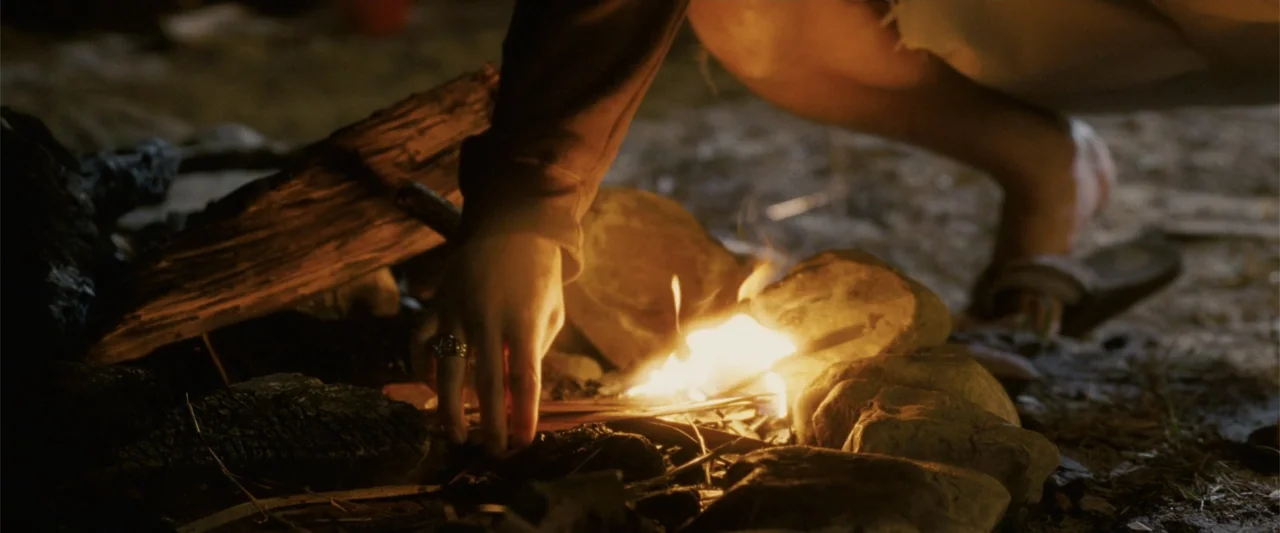 Dear John (2010) movie still: closeup — A dark outdoor space features a small, active campfire contained by smooth rocks. A person in dark long…