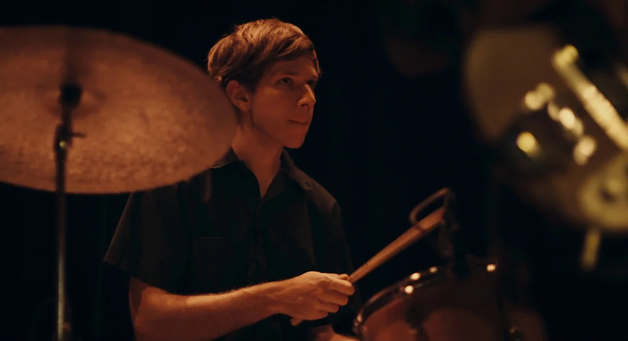 Mistress America (2015) movie still: medium — A dimly lit room focuses on a musician behind a drum set. The drummer stands ready with a drumstick in…