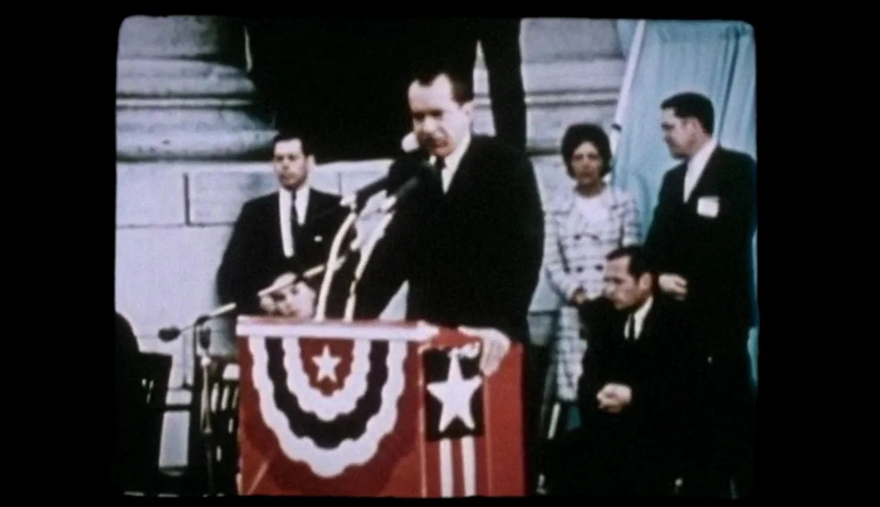 13th (2016) movie still: medium — A man stands behind a red podium decorated with patriotic bunting and stars. Several people sit and stand in the…
