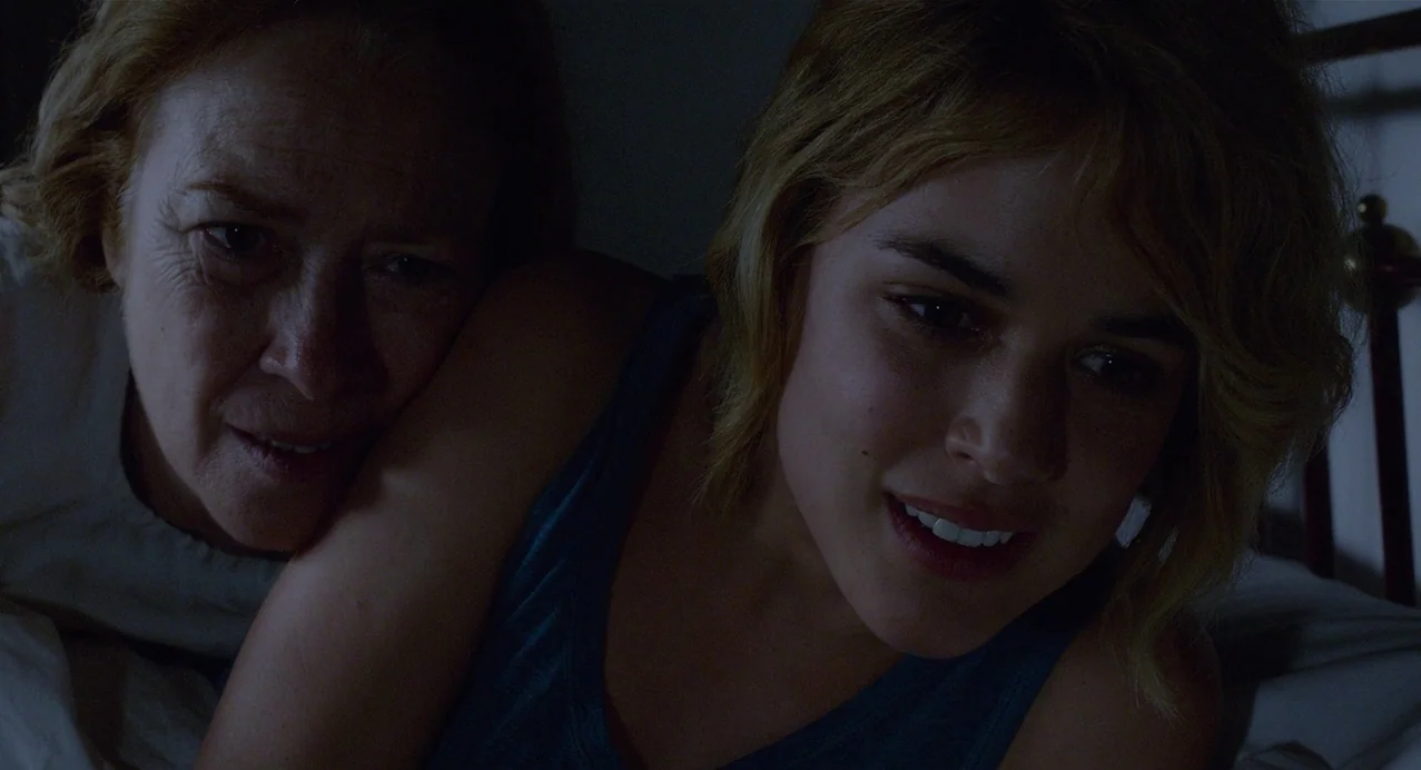 Julieta (2016) movie still: medium — Two women share a close space in a dark room under white bed sheets. The younger woman watches a screen while…