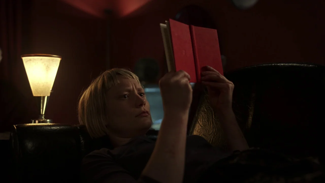 Piercing (2018) movie still: medium — A dimly lit room with deep red walls and dark furniture. A woman with blonde hair reads a small red book.