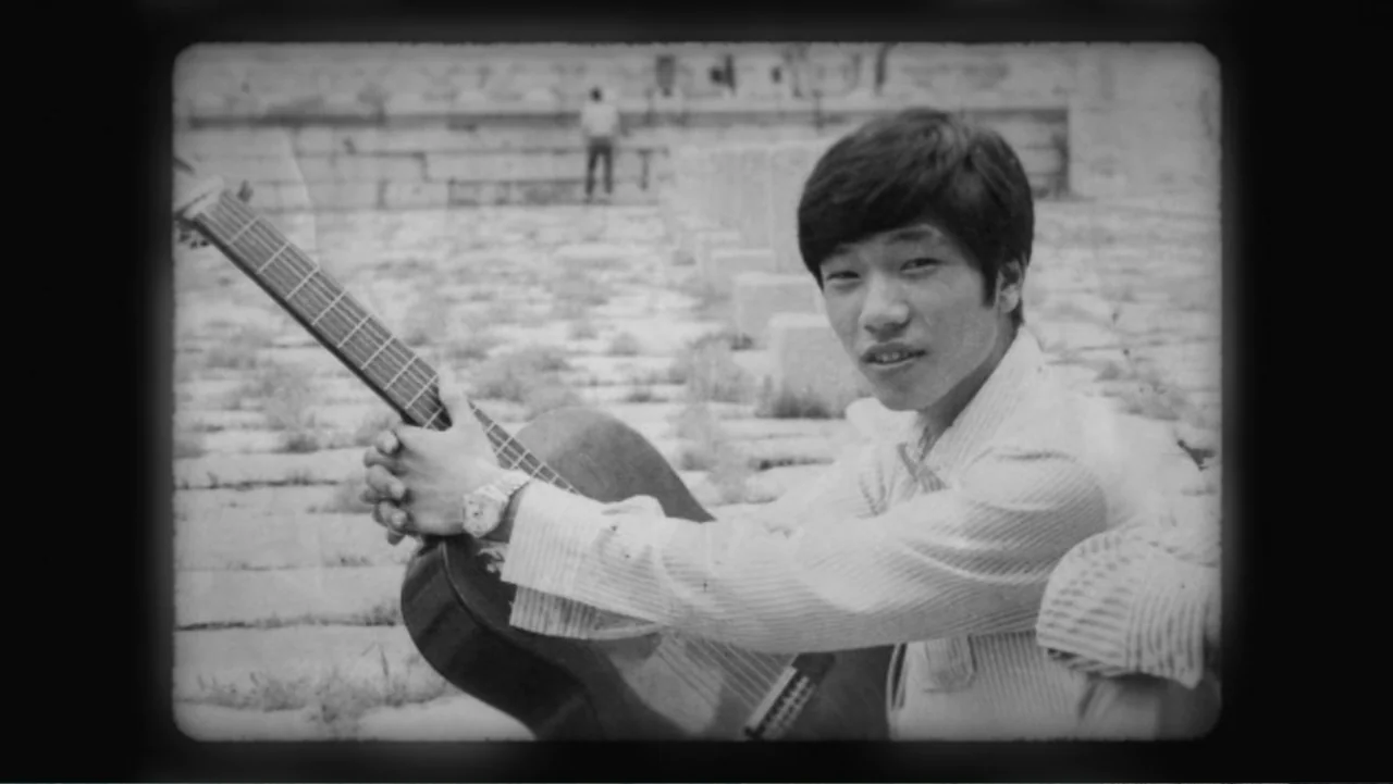 Lee Soo Man: King of K-Pop (2025) movie still: medium — A stone paved plaza features a distant figure standing against a low masonry wall. A young…
