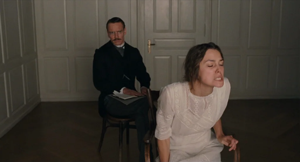 A Dangerous Method movie still: medium — A stark room with wainscoted white walls and a polished herringbone wood floor. A woman in white sits in a…