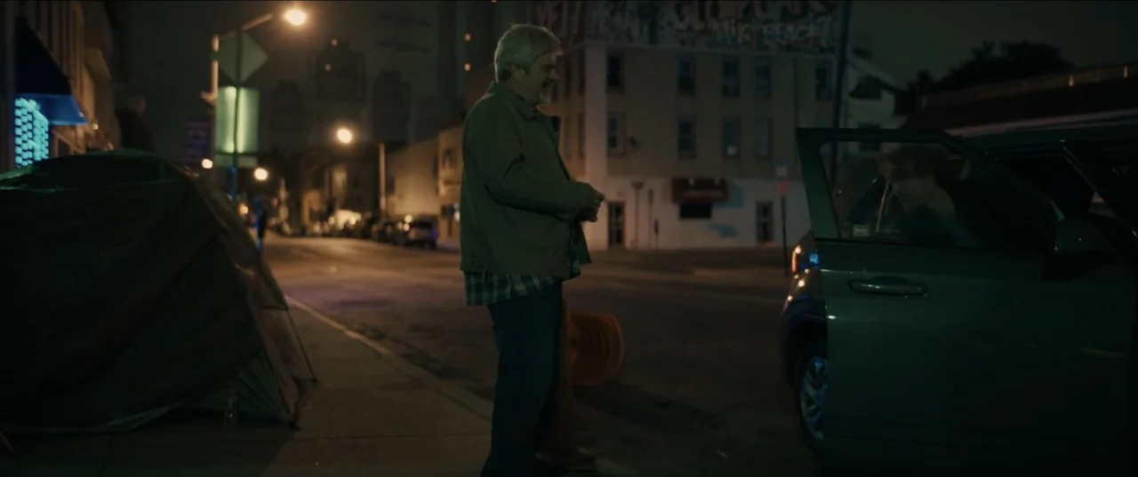 Guns Up (2025) movie still: fullbody — A nighttime city street features a parked car and a small tent on the sidewalk. An older man stands near the…