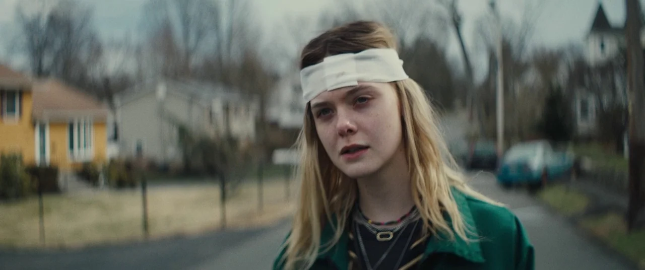 I Think We’re Alone Now (2018) movie still: medium — A quiet suburban street with houses and parked cars sits under an overcast sky. A young woman…