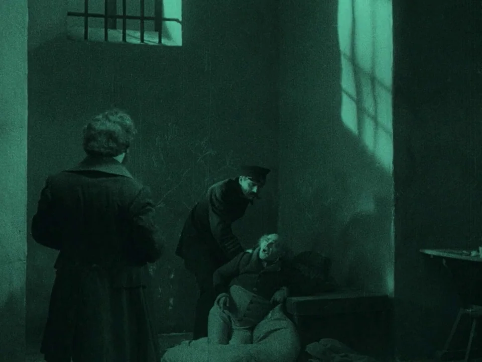 Nosferatu (1922) movie still: medium — A dark, spartan masonry cell features a high, barred window and stone walls. An officer leans toward a man…