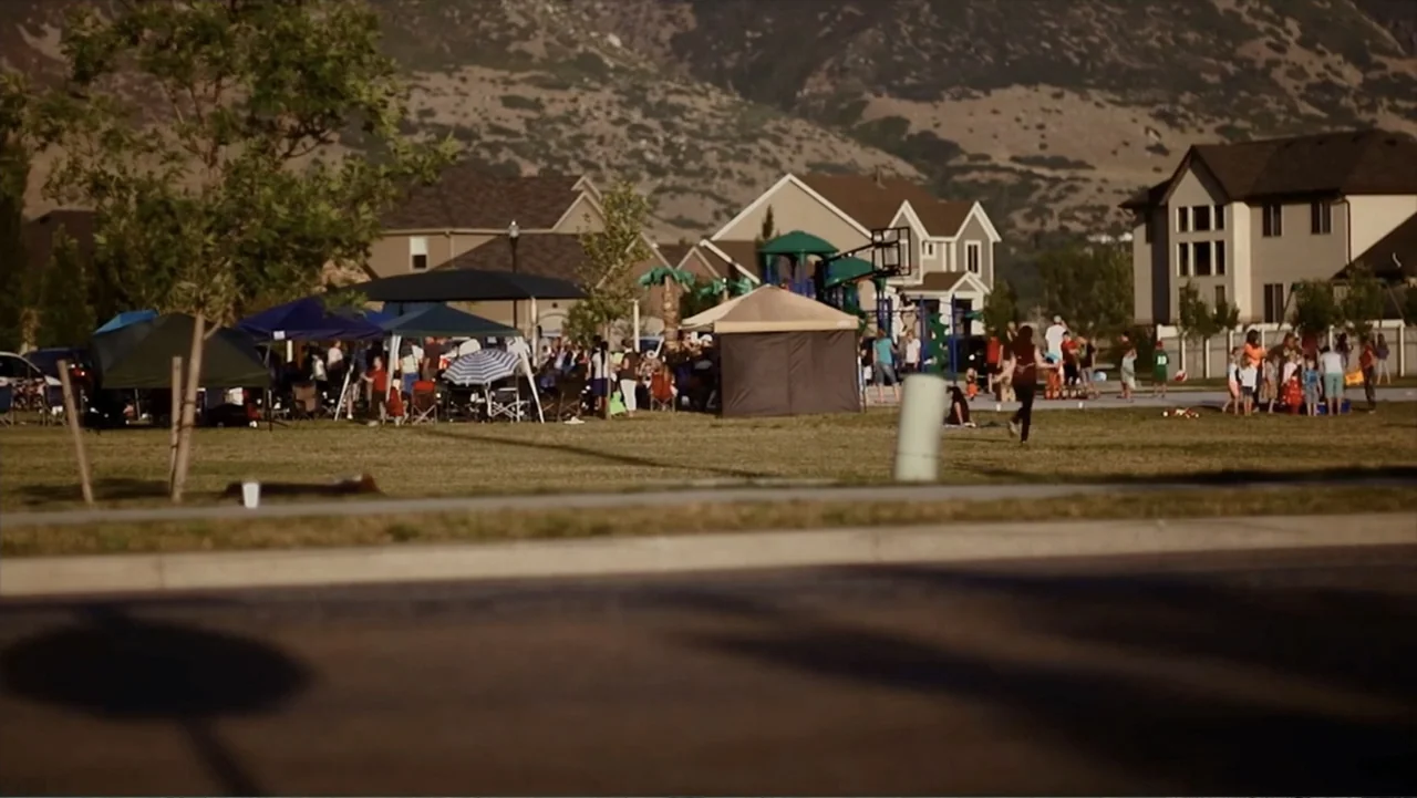 Hello I Love You (2018) movie still: wide — A community gathering occurs in a neighborhood park near residential houses and mountains. Numerous…