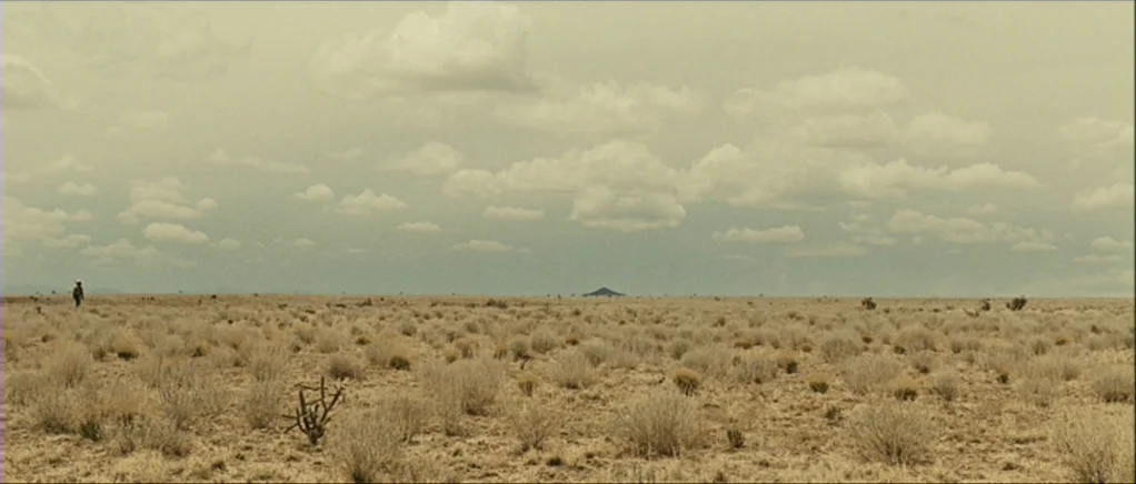 No Country For Old Men movie still: establishing — A vast, flat desert basin is covered in sparse, dried-out scrub and desert vegetation. A lone…