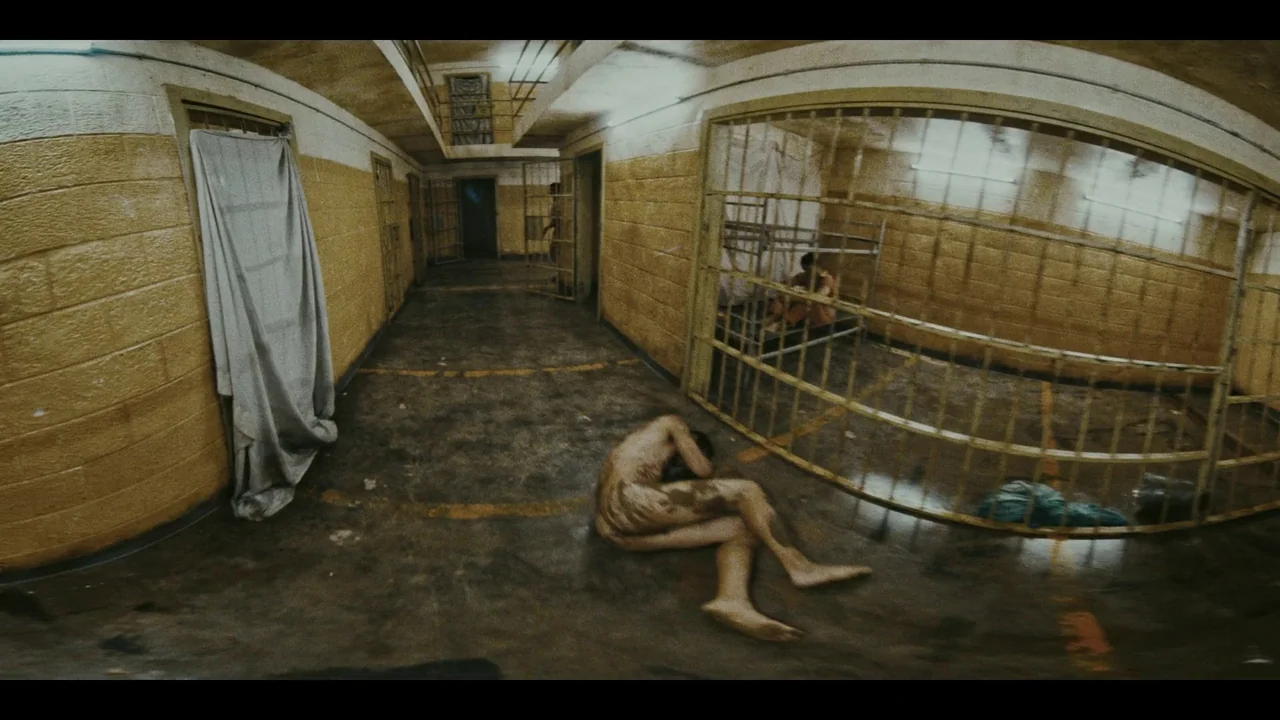 The Card Counter (2021) movie still: wide — A grimy corridor features yellow cinder block walls and metal cell bars on one side. A naked man lies…