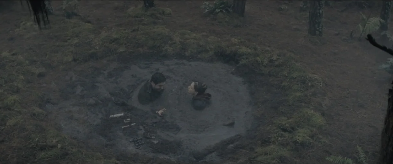 Quicksand (2023) movie still: wide — A deep circular pit of wet gray mud is located in a pine forest. A man and a woman are sitting partially…
