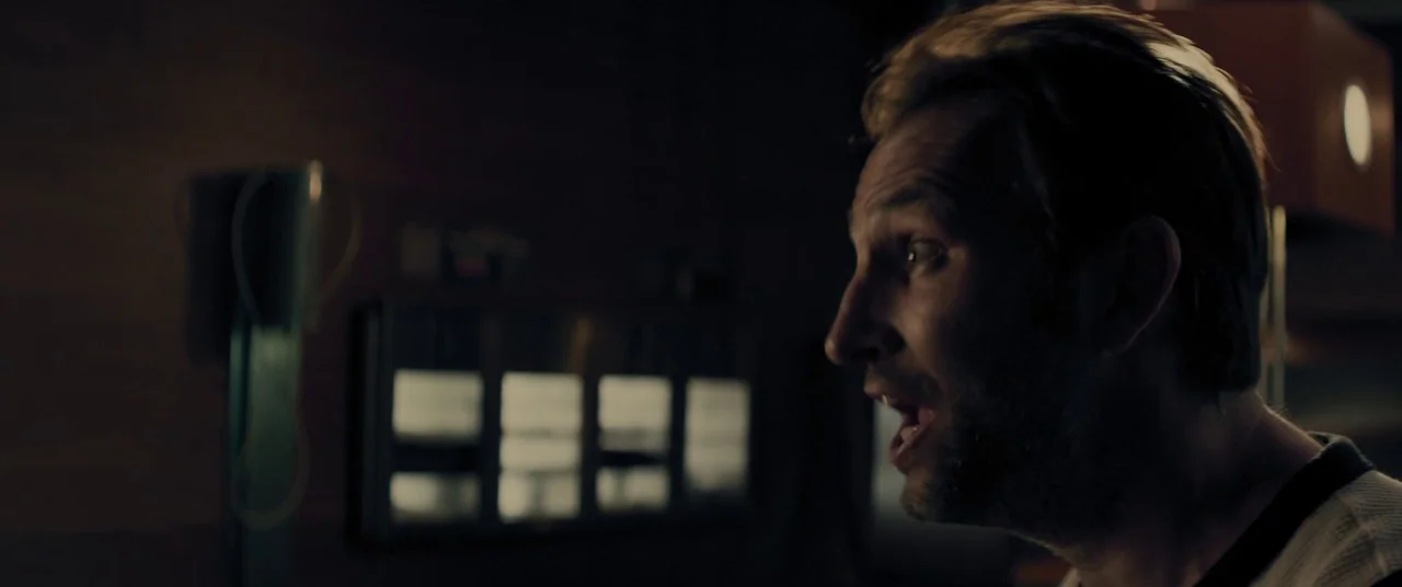 Domain (2017) movie still: closeup — A dimly lit interior space with blurry rectangular light fixtures visible in the background. A man stands in…