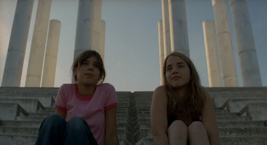 Water Lillies (2007) movie still: medium — Two teenage girls sit on concrete stairs in front of large, vertical stone pillars. The sky is a pale…