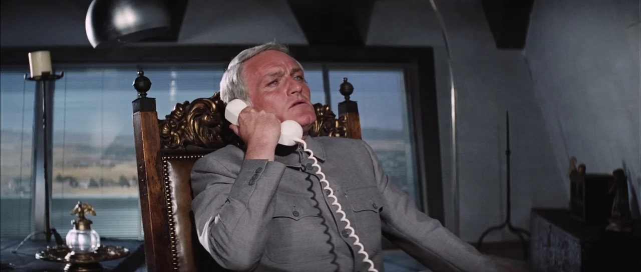 Diamonds Are Forever (1971) movie still: medium — A man sits in an ornate wooden chair inside a high-rise office space. He holds a white rotary…