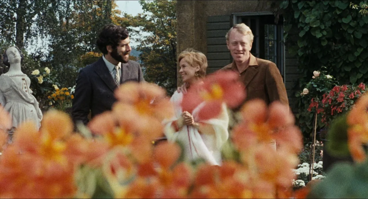 The Touch (1971) movie still: medium — Three people stand on a stone patio surrounded by vibrant orange flowers and greenery. The woman stands…
