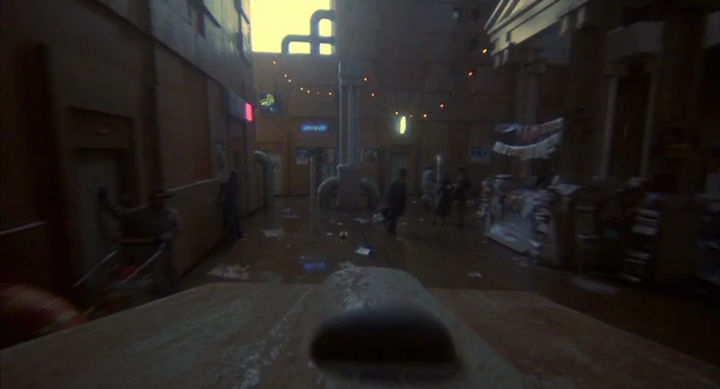 Brazil (1985) movie still: wide — A dimly lit pedestrian walkway is flanked by industrial-looking buildings at night. Figures traverse the wet…