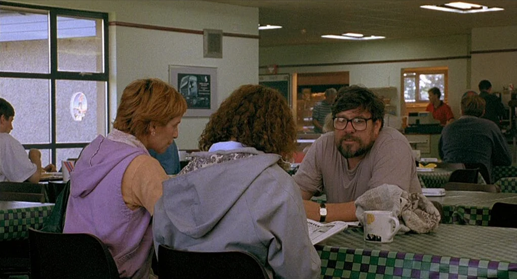 Butterfly Kiss (1995) movie still: medium — A utilitarian cafeteria features green and black checkered tablecloths and industrial fluorescent…