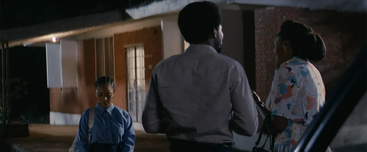 Ms. Kanyin (2025) movie still: medium — A night scene outside a brick motel building with lit walkways. Three people stand near each other in a…