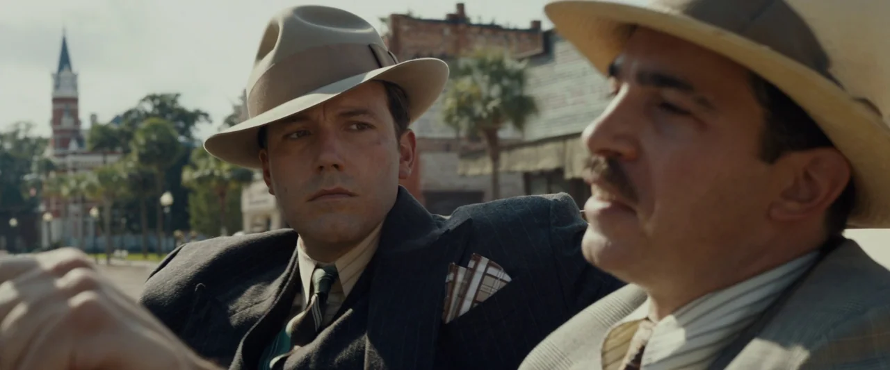 Live By Night (2016) movie still: medium — A sunny town square with old-fashioned buildings and palm trees in the background. Two men in fedoras sit…