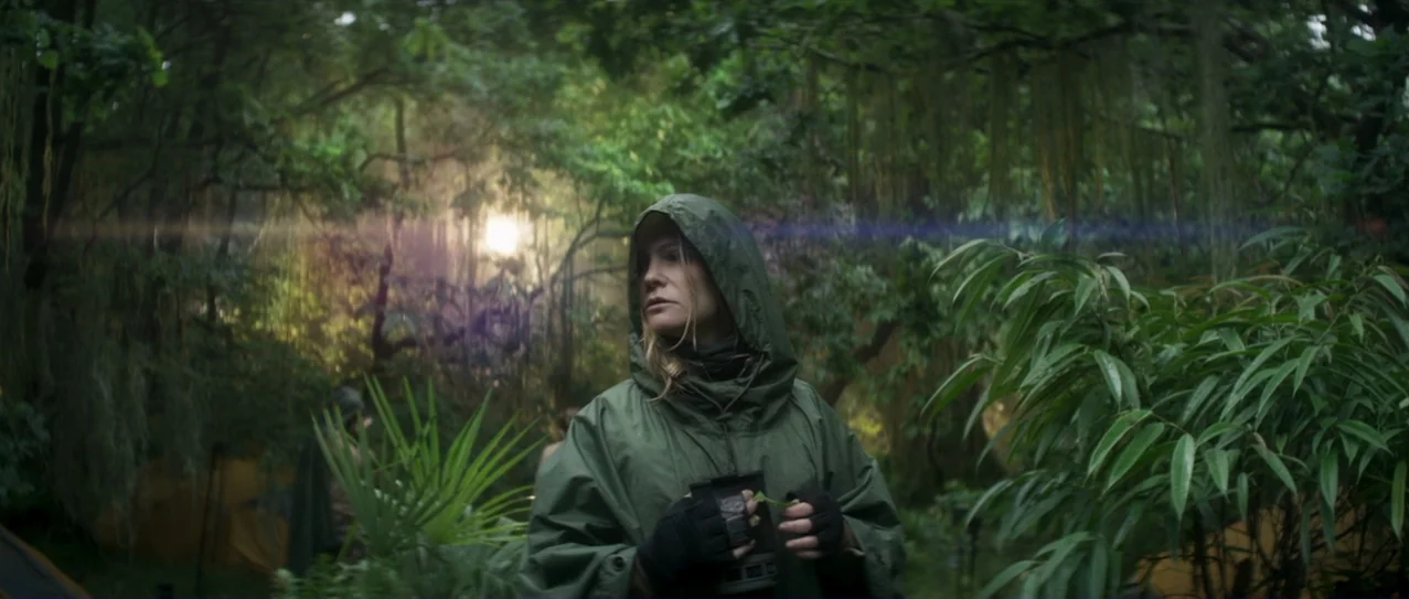 Annihilation (2018) movie still: medium — A dense, humid tropical forest floor features lush foliage and hanging mossy vines. A woman in a green…