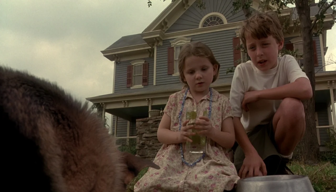 Signs (2002) movie still: medium — A suburban house with gray horizontal siding sits behind a grassy lawn. A young boy and a girl kneel on the…
