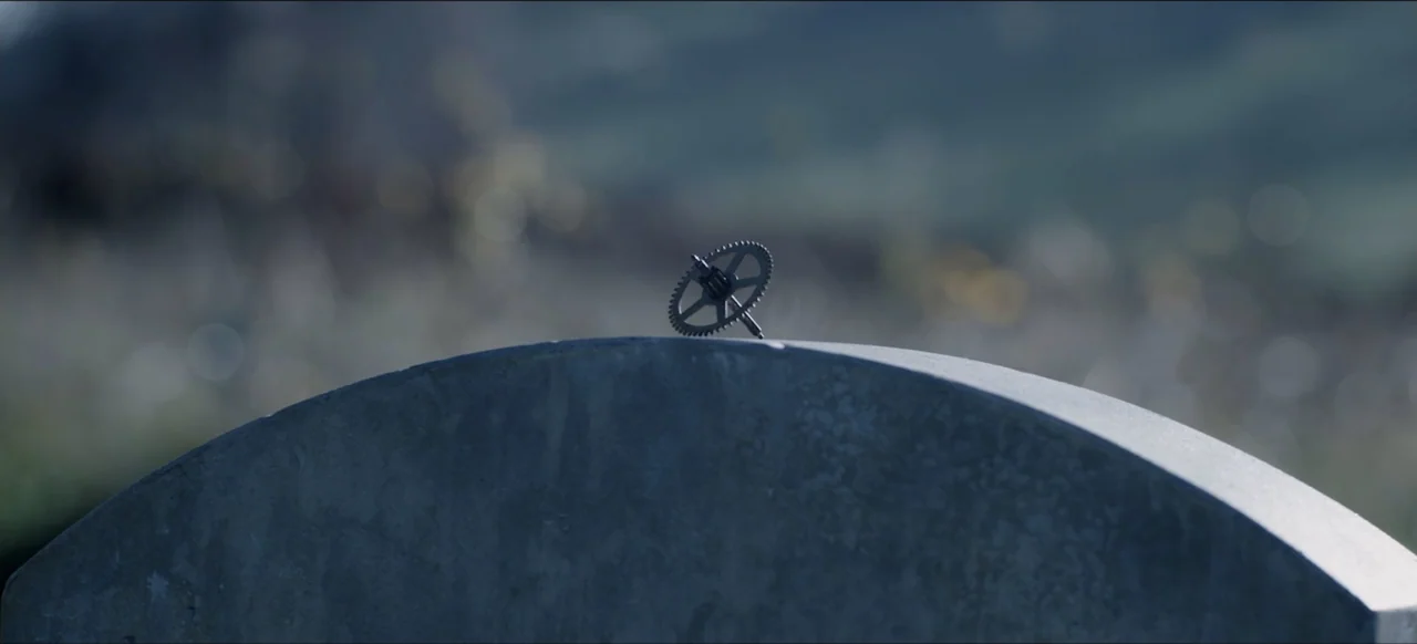We Go In at Dawn (2020) movie still: closeup — A stone headstone stands in the foreground against a blurred outdoor grassy field background. A small…