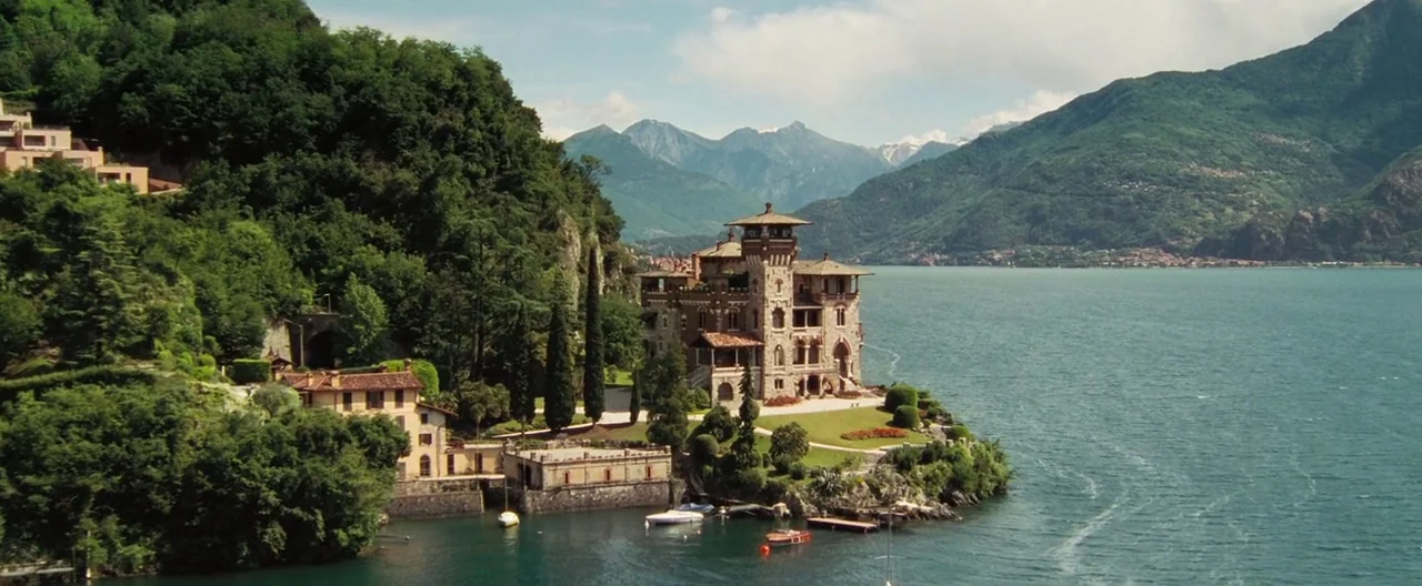 Casino Royale (2006) movie still: establishing — A stone villa sits on a lush, forested peninsula protruding into a large lake. Mountains rise in…