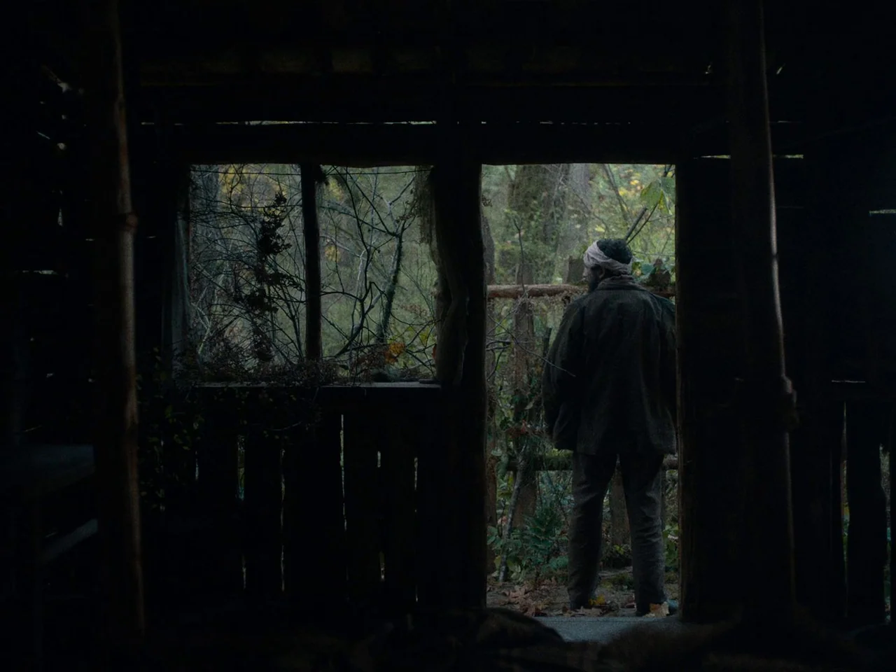 First Cow (2019) movie still: fullbody — A dark, rustic wooden structure with a large open threshold leading into a forest. A man stands in the…