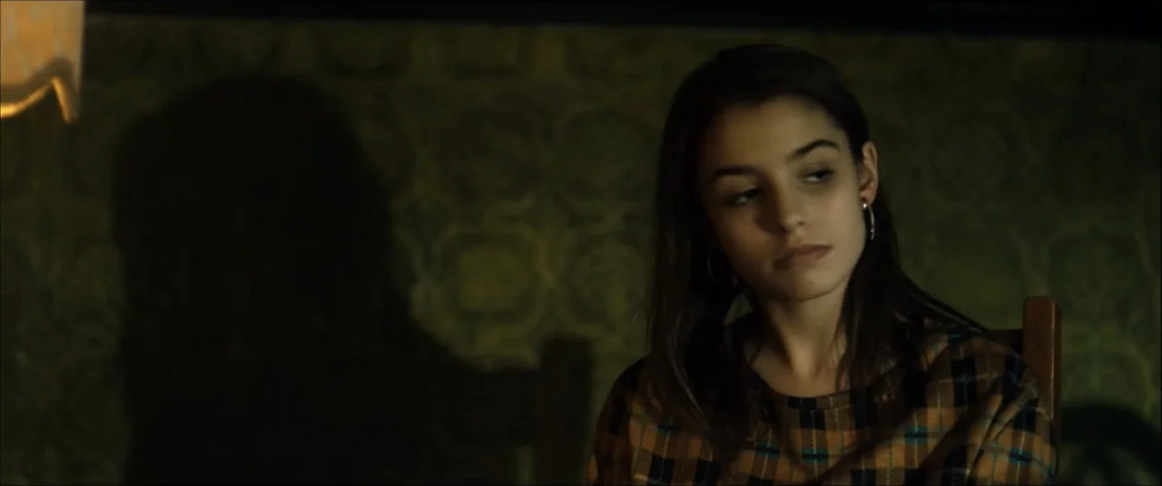 I'll Take Your Dead (2018) movie still: medium — A dimly lit room features a dark wall covered in patterned, muted green wallpaper. A young woman in…