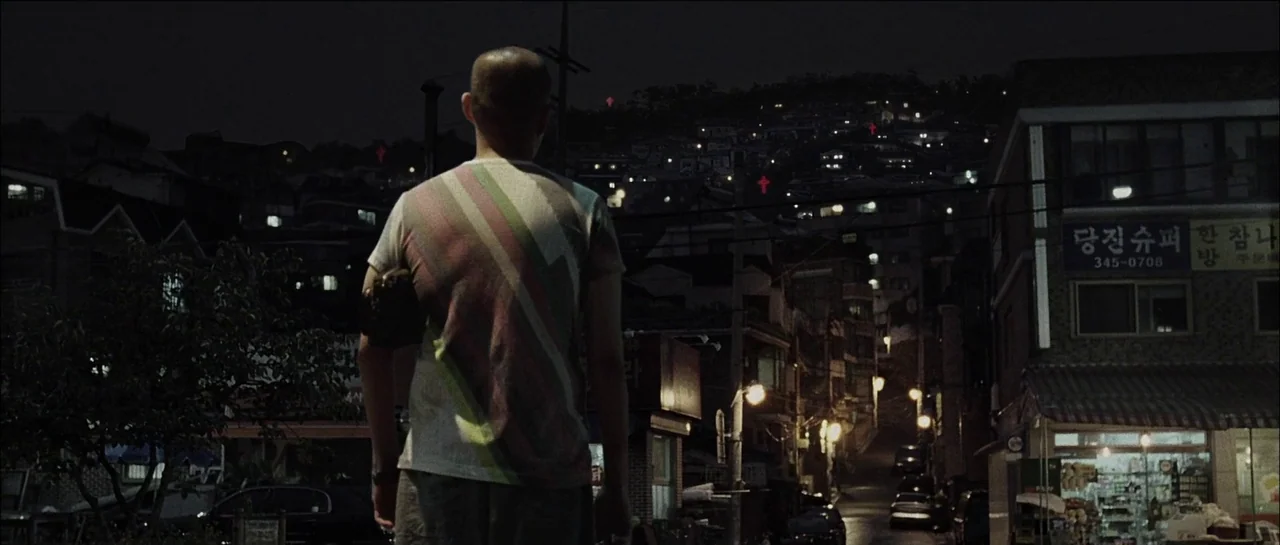 The Chaser (2008) movie still: medium — A man stands on a road at night in front of a hillside neighborhood. Numerous red neon crosses glow on the…