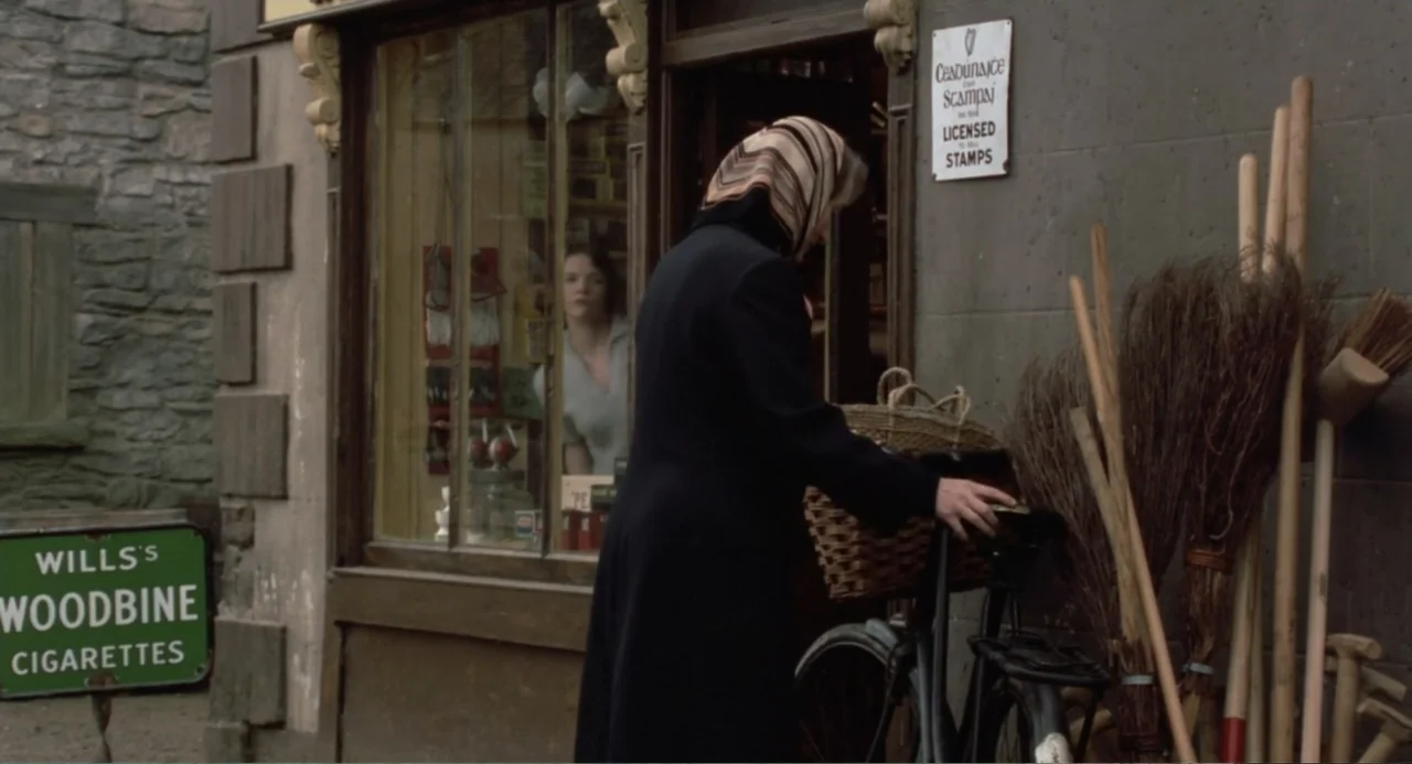 Dancing at Lughnasa (1998) movie still: medium — A rural shop front features stone walls, a display window, and brooms resting against siding. A…