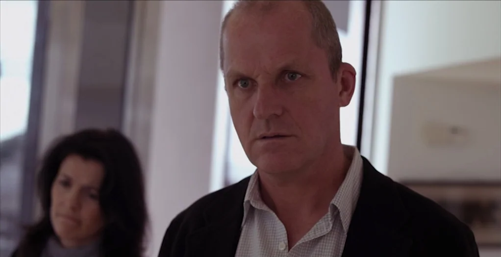 Dollhouse (2012) movie still: medium — A man and a woman stand in a modern, bright office interior. The man is positioned in the foreground while…