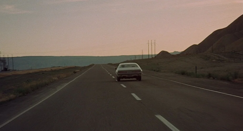 Vanishing Point (1971) movie still: wide — A desolate paved road cuts through a vast, arid landscape during golden hour. A white muscle car drives…