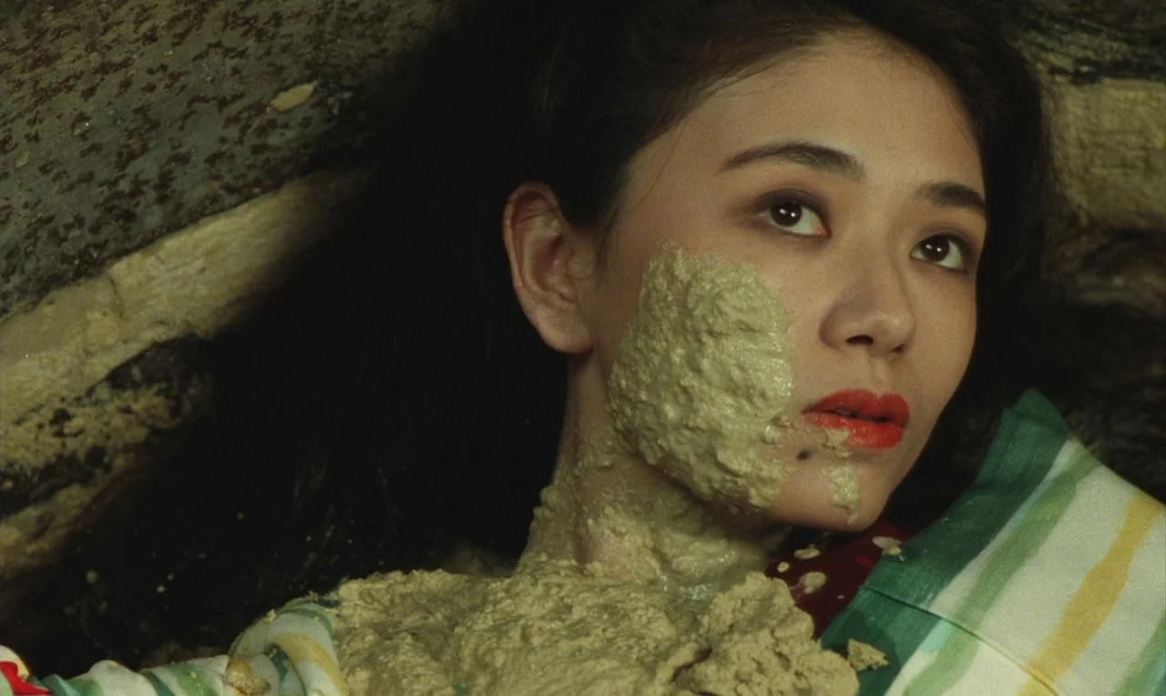 Yumeji (1991) movie still: closeup — A dark, cramped space with rough, aged stonework visible in the background. A woman lies with thick, pale green…