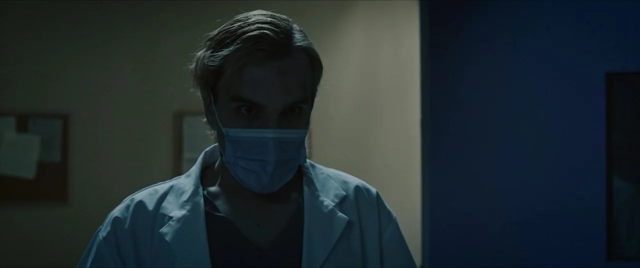 Vicious Fun (2021) movie still: medium — A sterile medical hallway with blue walls and two bulletin boards in the background. A doctor in a white…
