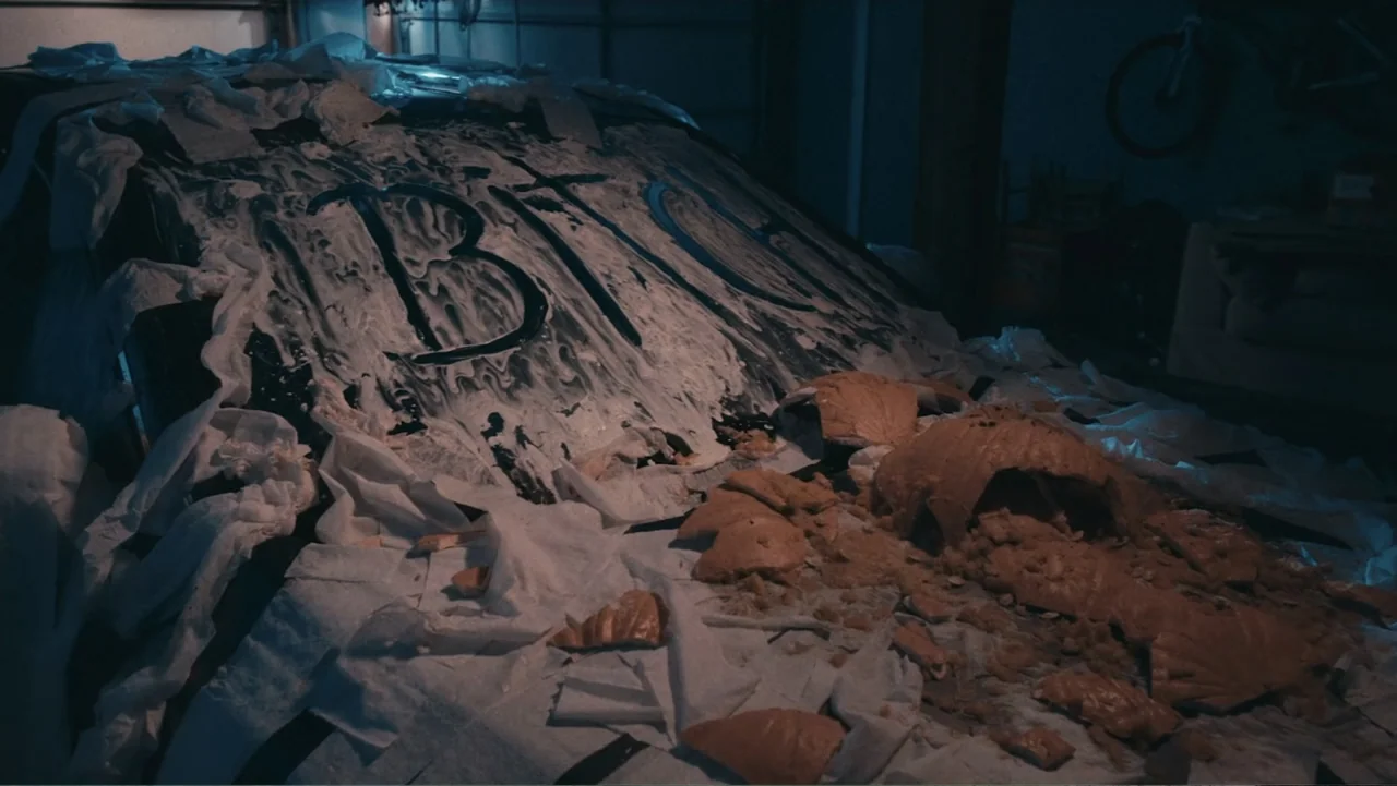Terrifier 2 (2022) movie still: medium — A dark garage interior shows the hood of a car covered in white paper strips. A smashed orange pumpkin lies…