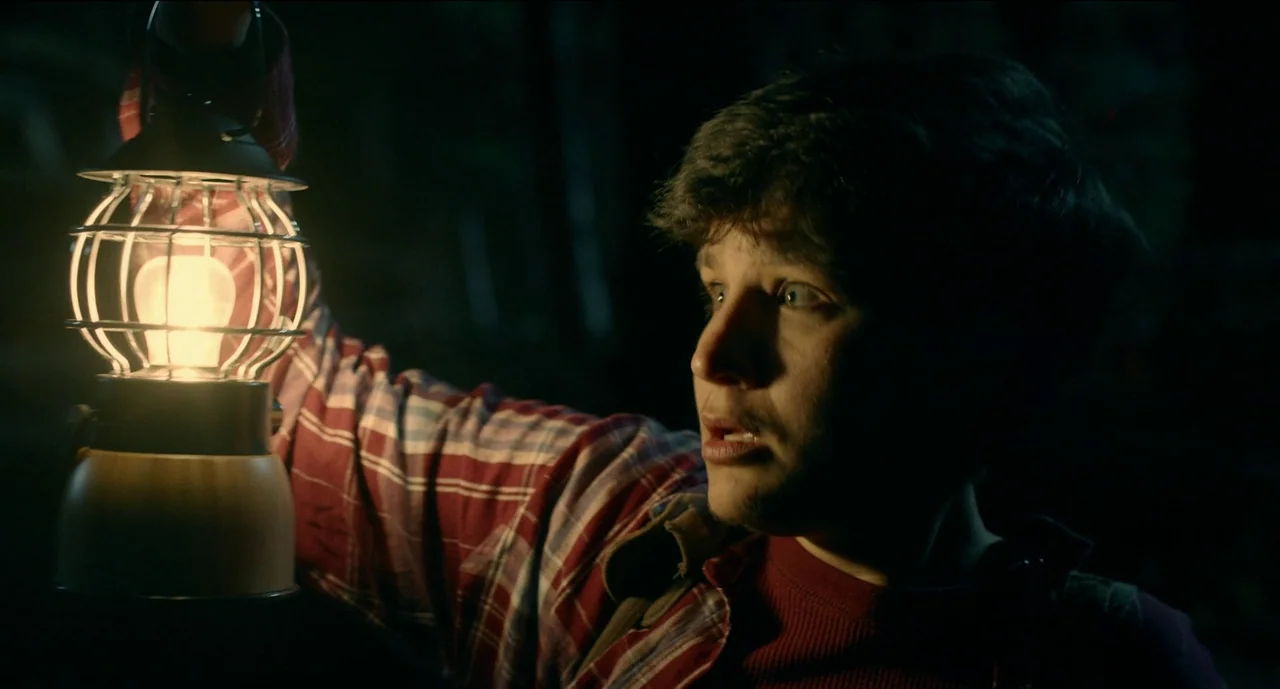 Trim Season (2023) movie still: closeup — A dark outdoor setting surrounded by deep shadows in the night. A young man illuminates the space with a…