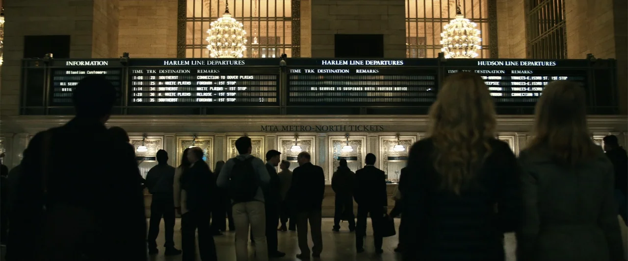 21 Bridges (2019) movie still: wide — A grand terminal lobby features ornate ceiling lights and a large digital departure board. Numerous people…
