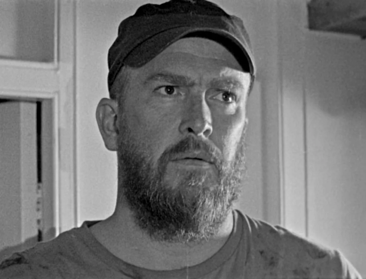 Bait (2019) movie still: closeup — A man stands indoors against a plain wall with visible molding. He wears a baseball cap and looks toward the…