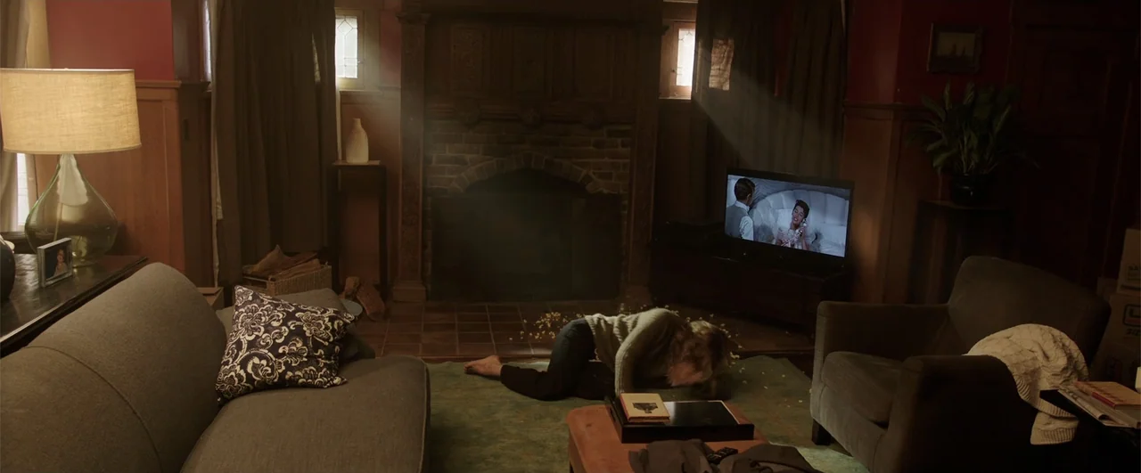 Lights Out (2016) movie still: wide — A dark living room contains a brick fireplace, upholstered furniture, and a television screen. A woman crawls…