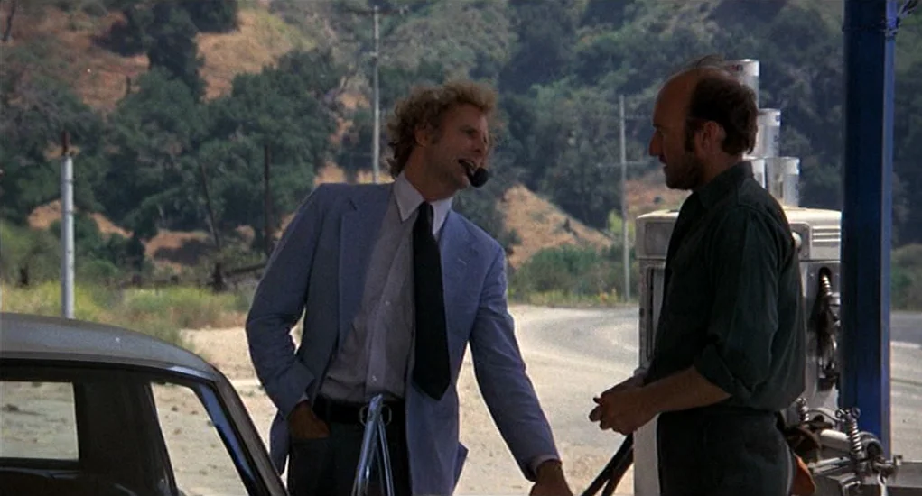 Family Plot (1976) movie still: medium — A man in a light blue suit and a man in a dark shirt talk at a rural gas station. Rolling hills and power…