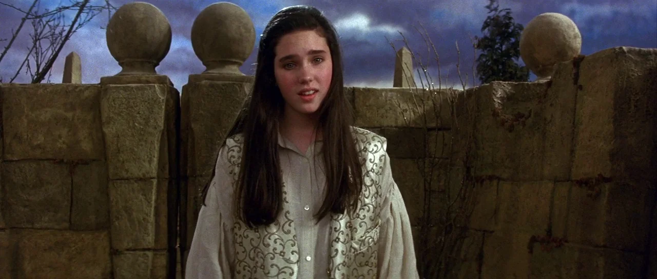Labyrinth (1986) movie still: medium — A young woman stands in front of a heavy weathered stone wall at dusk. The sky behind her is filled with…