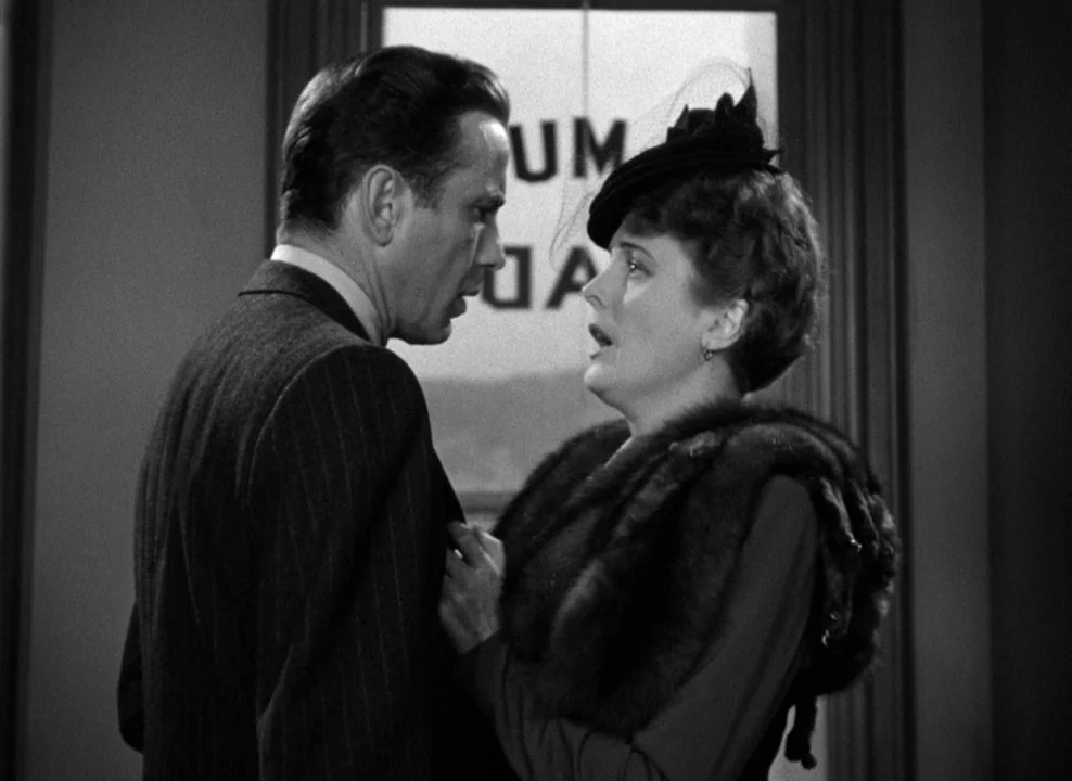The Maltese Falcon (1941) movie still: medium — An interior office with a glass-paneled door in the background featuring partial text lettering. A…