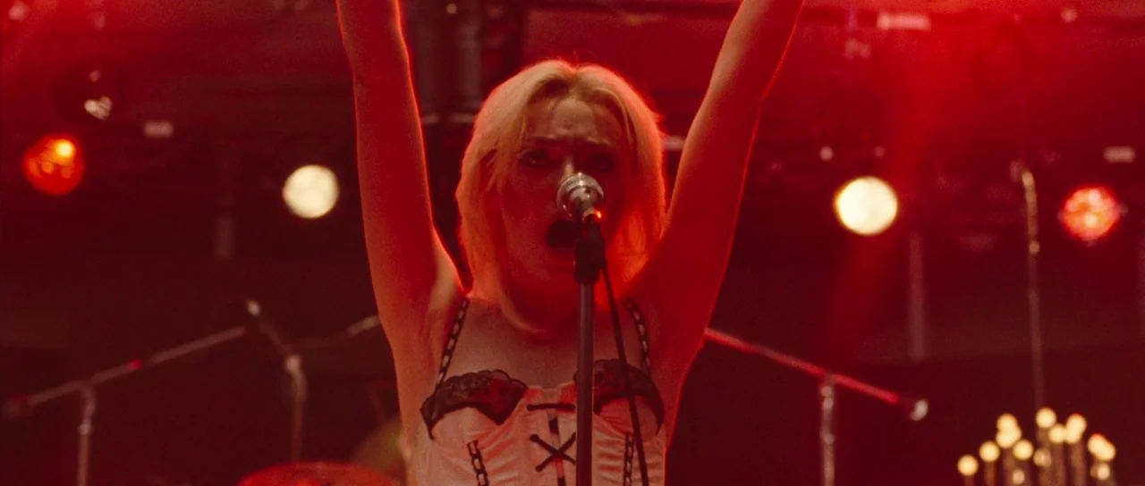 The Runaways (2010) movie still: medium — A dimly lit concert venue with heavy red stage lighting and blurred instrument silhouettes. A blonde woman…